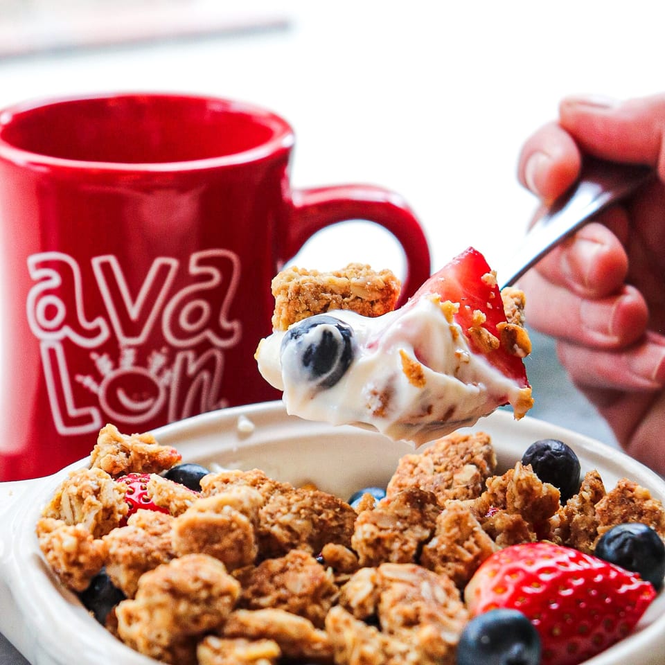 Bowl of yogurt topped with granola, strawberries, and blueberries, with a spoonful lifted beside a red coffee mug.