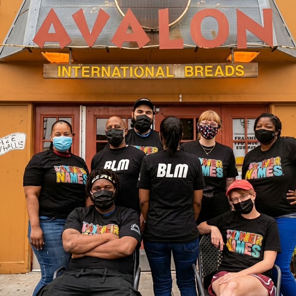 Eight diverse people in masks and "Power to the Names" or "BLM" shirts stand outside Avalon International Breads.