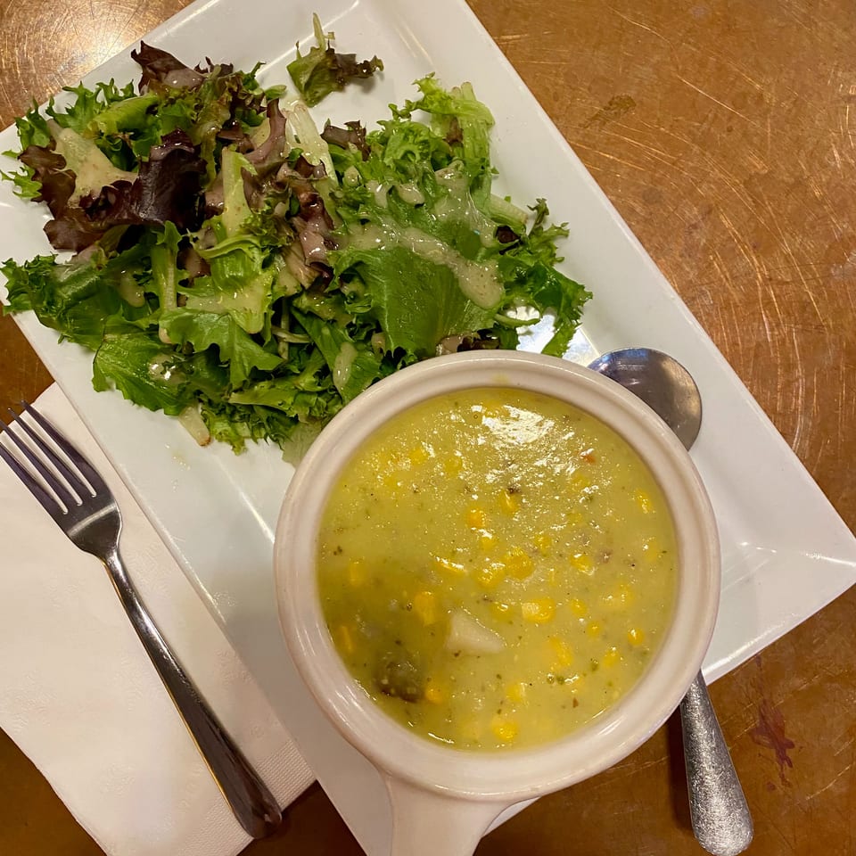 Mixed green salad with creamy dressing and a bowl of corn chowder served on a white plate with utensils.