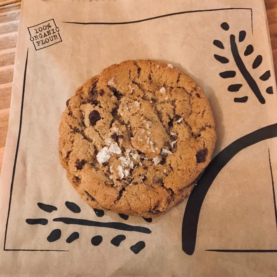 Large chocolate chip cookie sprinkled with flaky salt on branded brown bakery paper marked 100% organic flour.