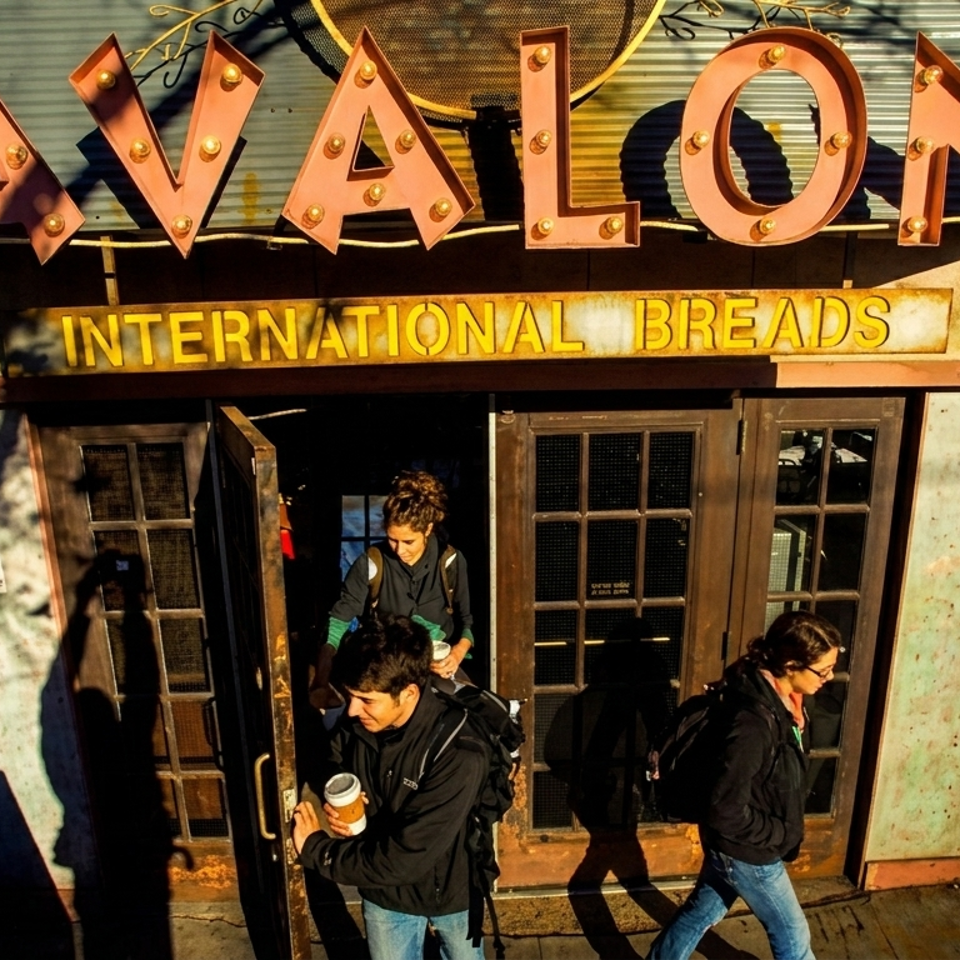 People exit Avalon International Breads, a brick building with a large illuminated sign, in warm sunlight.